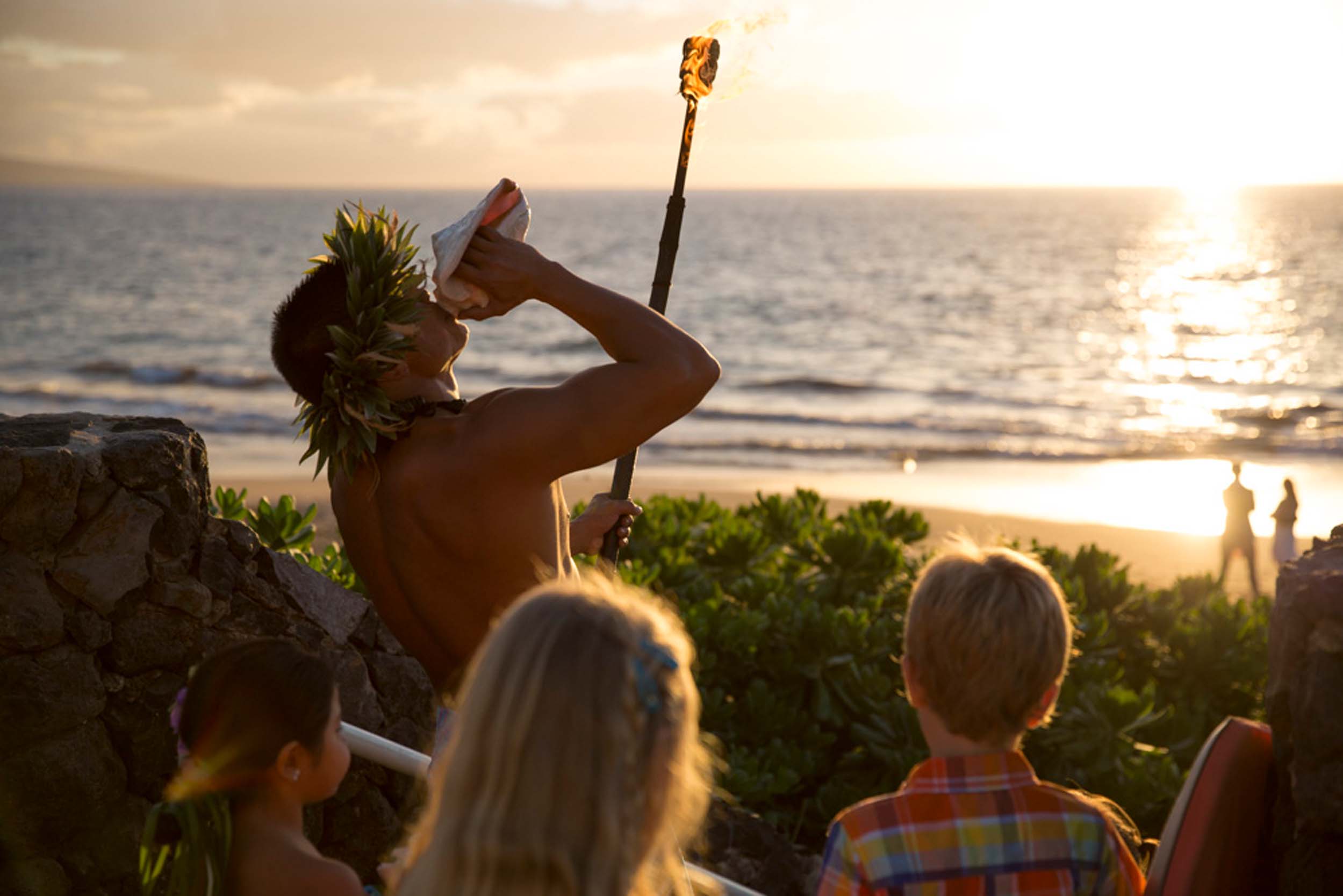 a group of kits watching the sunset as a man blows into a shell